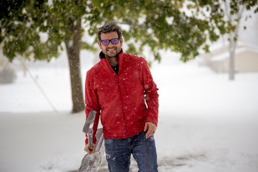 A male wearing a red jacket and holding a snow shovel while smiling to the camera with a blurred background