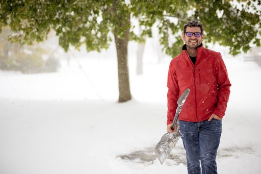 A closeup shot of a male wearing a red jacket and holding a snow shovel while smiling and looking at the camera