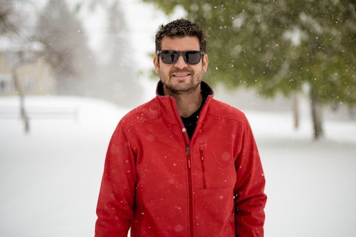 A closeup shot of a male wearing a red jacket and smiling while looking at the camera with a blurred background