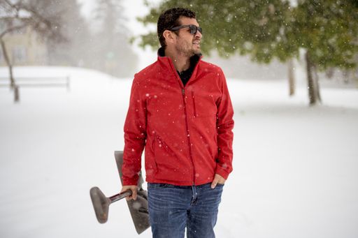 A closeup shot of a male wearing a red jacket and holding a snow shovel with his hand in the pocket