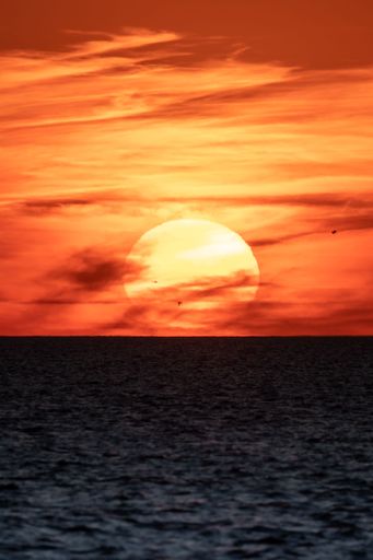 A vertical of a big glowing sun captured in the red sunset sky above a tranquil evening seascape
