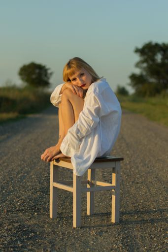A vertical shot of a beautiful caucasian woman wearing a white shirt, sitting on a stool outdoors next to a field