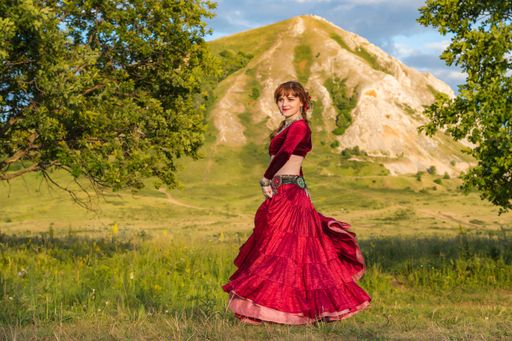 Young beautiful red-headed girl in red ball gown is dancing incendiary dance on green meadow against backdrop of mountain and summer sunset.