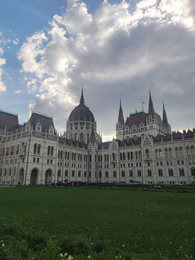 A vertical shot of the Parliament building in Budapest, Hungary with a park and a cloudy sky in the background.