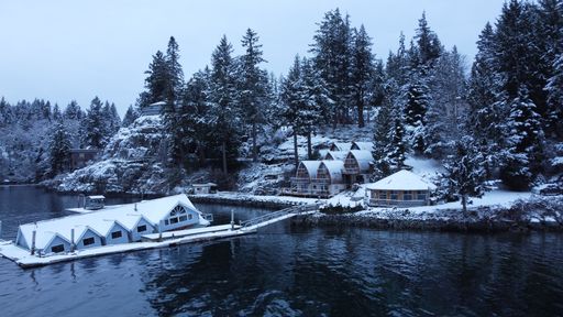 A drone shot of a lakefront with cabins covered in the snow surrounded by coniferous trees