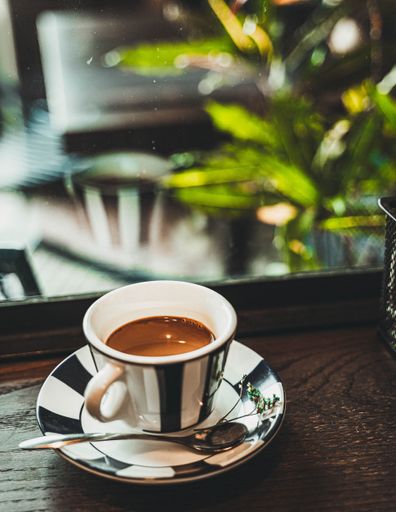 A vertical closeup of coffee in the little black and white striped cup, with a spoon near on the wooden table plants and floor blurred background