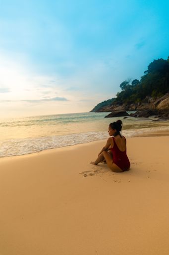 A vertical shot of a female wearing a red swimsuit sitting at the sandy beach during sunrise