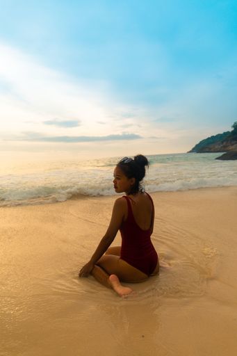 A vertical shot of a female wearing a red swimsuit sitting at the sandy beach during sunrise