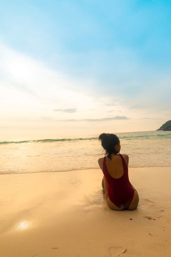 A vertical shot of a female wearing a red swimsuit sitting at the sandy beach during sunrise