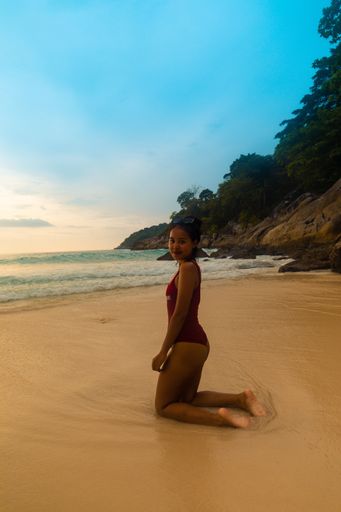 A vertical shot of a female wearing a red swimsuit sitting at the sandy beach during sunrise