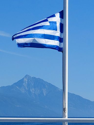 A vertical closeup shot of the Greece flag on the edge of a boat with a mountain in the background