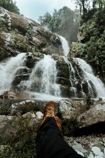 A vertical shot of a feet and shoes of a man with a scenic waterfall on the background