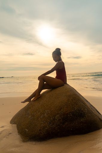 A vertical shot of an attractive female wearing a red swimsuit sitting on a rock at the beach