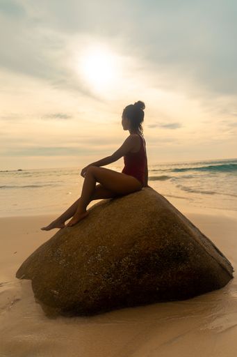 A vertical shot of an attractive female wearing a red swimsuit sitting on a rock at the beach