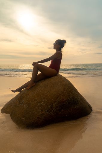 A vertical shot of an attractive female wearing a red swimsuit sitting on a rock at the beach