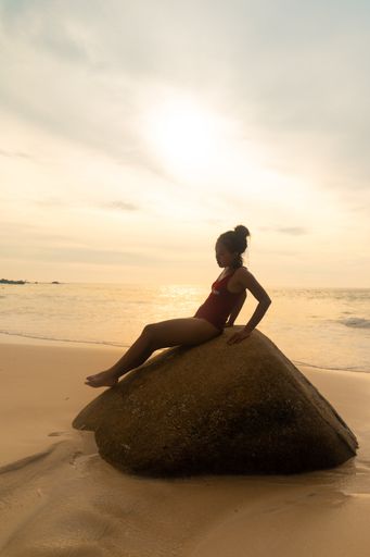 A vertical shot of an attractive female wearing a red swimsuit sitting on a rock at the beach
