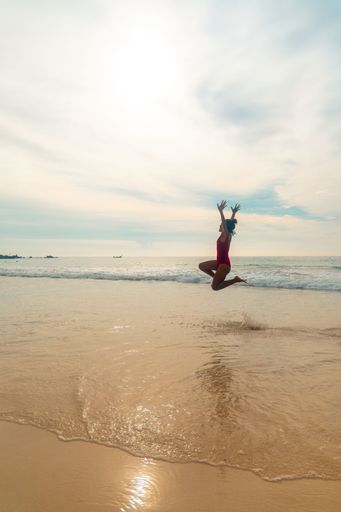 A vertical shot of a female wearing a red swimsuit happily jumping at the beach enjoying the nice weather