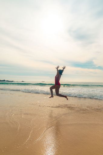 A vertical shot of a female wearing a red swimsuit happily jumping at the beach enjoying the nice weather