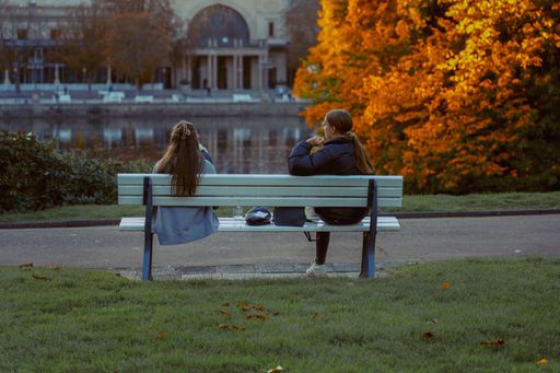 A scenic view of two girls sitting on a bench in the park with a background of a pool and autumn trees
