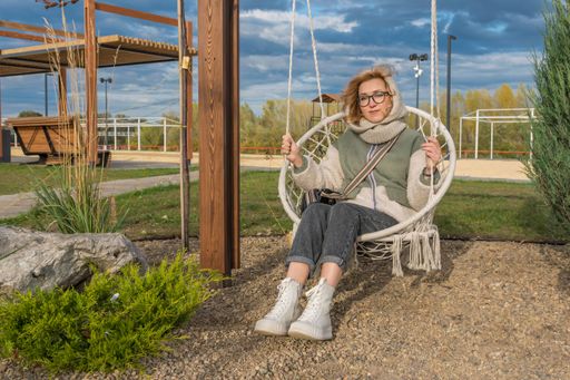 Young blonde girl in glasses and insulated autumn clothes rides on swing in recreation and amusement park against of landscape. Looks at camera.