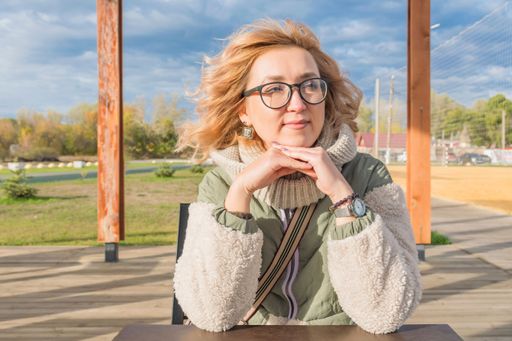 Young pretty blonde girl in warm autumn clothes sits in gazebo outdoors. Look away. Hair blowing in wind.