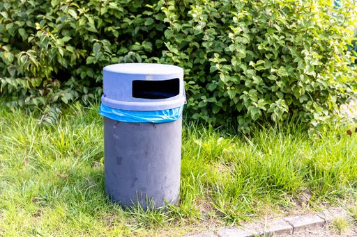 A closeup of a metallic garbage bin in a field covered in greenery under the sunlight