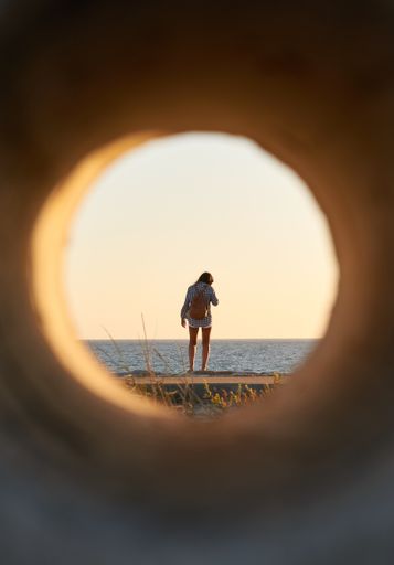 A vertical shot of a woman standing on the beach seen through a pipe