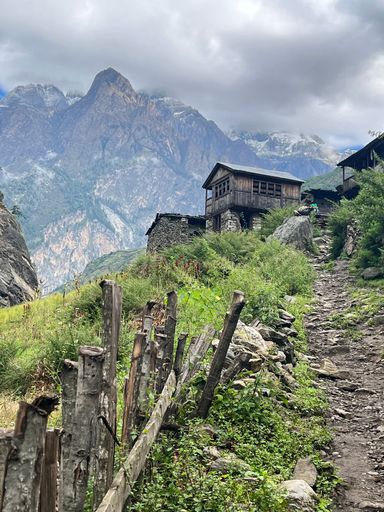 A rocky path with a wooden cabin and mountains under a cloudy sky in the background.