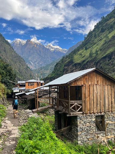 A vertical shot of a wooden cabin and a person hiking with mountains and a cloudy sky in the background.