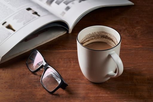 A glass of ground coffee milk, reading glasses and a magazine on a wooden table