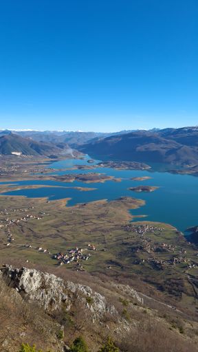 A vertical shot of a blue lake, shoreline buildings and mountains under the clear blue sky