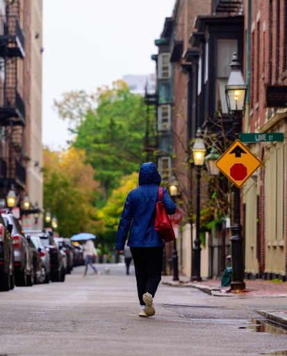 A back shot of female in a blue raincoat walking between building in a City of Boston During the Fall