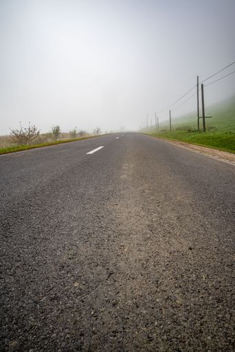 A vertical shot of a beautiful road with green fields on the sides with cloudy and foggy sky in background