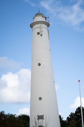 A vertical shot of a white lighthouse with trees and a cloudy sky in the background.