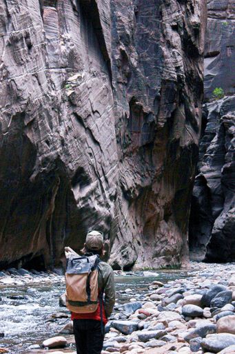 A vertical shot of a backpacker standing on a creek shore and observing the rocky walls of a canyon