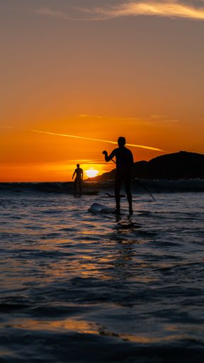 A vertical shot of tow surfers during sunset with the sun in the background