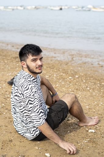 A Hispanic man sitting on the shore with the view of the sea in the background