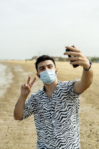 A Hispanic man wearing protective mask while taking selfies with peace hand sign at a beach