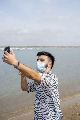 A Hispanic man wearing protective mask while taking selfies with peace hand sign at a beach