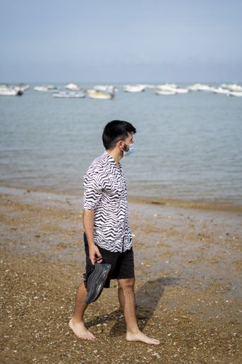 A Hispanic man wearing protective mask strolling barefooted at a beach on a sunny day